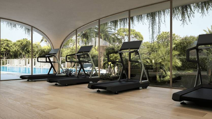Trois tapis roulants dans une salle de sport intérieure moderne avec de grandes fenêtres incurvées donnant sur une piscine extérieure tranquille entourée d'une verdure luxuriante, idéale pour un investissement à Dubaï.