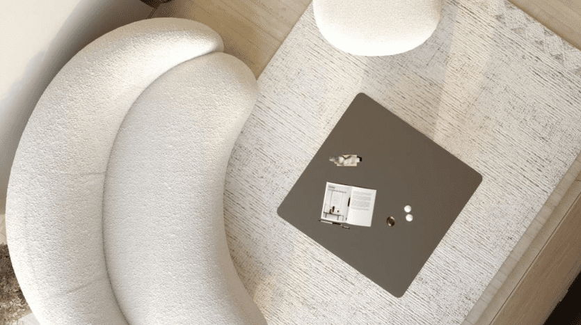 Vue aérienne d'un salon moderne dans un appartement de Dubaï avec un canapé blanc aux courbes uniques et une table basse carrée noire avec un livre et de petits objets dessus, posés sur un fond blanc cassé texturé