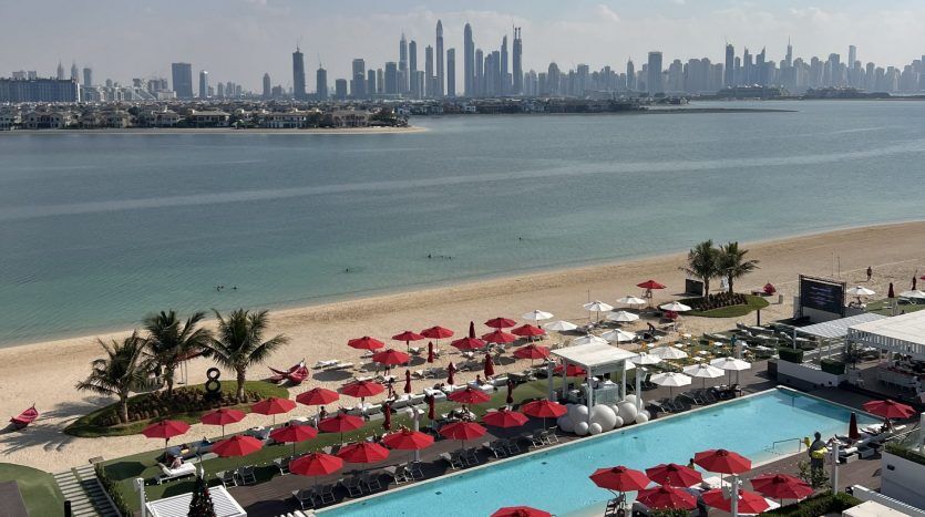 Vue d'un front de mer avec des parasols rouges, une piscine et des transats face à la mer avec un horizon de ville lointain sous un ciel clair, illustrant parfaitement une opportunité d'investissement exceptionnelle à Dubaï.