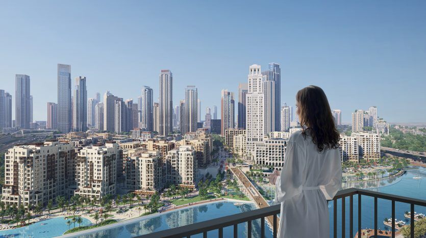 Une femme vêtue d'une robe blanche se tient sur un balcon surplombant l'horizon luxueux de Dubaï avec des gratte-ciel modernes et un canal sinueux, sous un ciel bleu clair.