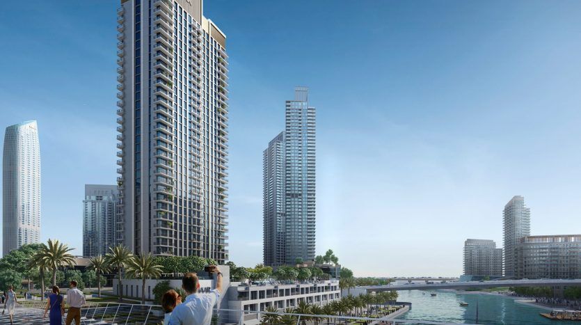Trois personnes observant des gratte-ciel modernes depuis une promenade luxuriante au bord de l'eau à Dubaï, mettant l'accent sur le développement urbain et les vues panoramiques.