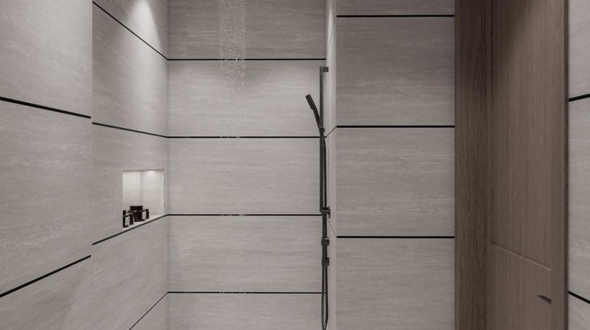 Une salle de bains moderne dans un appartement de Dubaï avec des murs et du sol carrelés gris, comprenant une cabine de douche en verre avec eau courante, une pomme de douche à main et des armoires en bois.