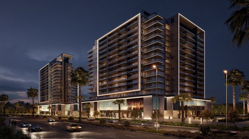 Vue nocturne d'un complexe hôtelier moderne à plusieurs étages avec des balcons éclairés, entouré de palmiers et de voitures passant dans la rue : idéal pour investir à Dubaï.