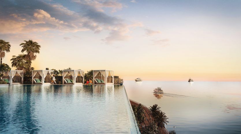 Une piscine à débordement surplombant une mer sereine au coucher du soleil, avec des palmiers et des cabines modernes semi-circulaires au bord de la piscine. Des bateaux sont visibles à l'horizon sous un ciel pastel près d'une luxueuse villa à