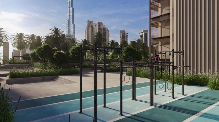 Une salle de sport en plein air avec divers équipements de fitness sur un tapis bleu, entourée de plantes vertes et à proximité d'immeubles de grande hauteur, dont une villa à Dubaï, avec un grand gratte-ciel en arrière-plan sous un ciel clair