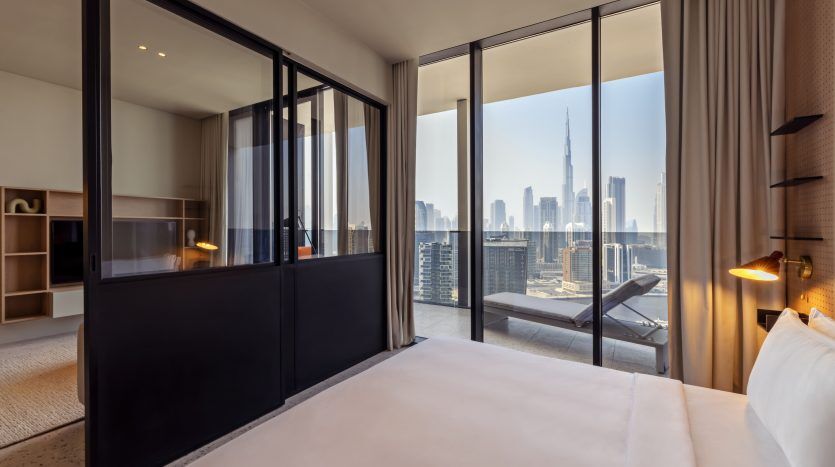 Une chambre d'hôtel moderne dotée d'un grand lit, d'une cloison vitrée et de grandes fenêtres offrant une vue panoramique sur les toits de Dubaï avec de grands immeubles sous un ciel dégagé.
