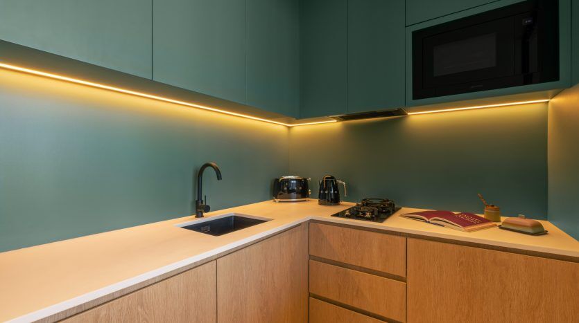 Kitchenette moderne dans un appartement de Dubaï avec murs bleu sarcelle, éclairage sous meuble et armoires en bois. Comprend des appareils électroménagers comme un grille-pain et une cuisinière, avec un livre ouvert sur le comptoir.