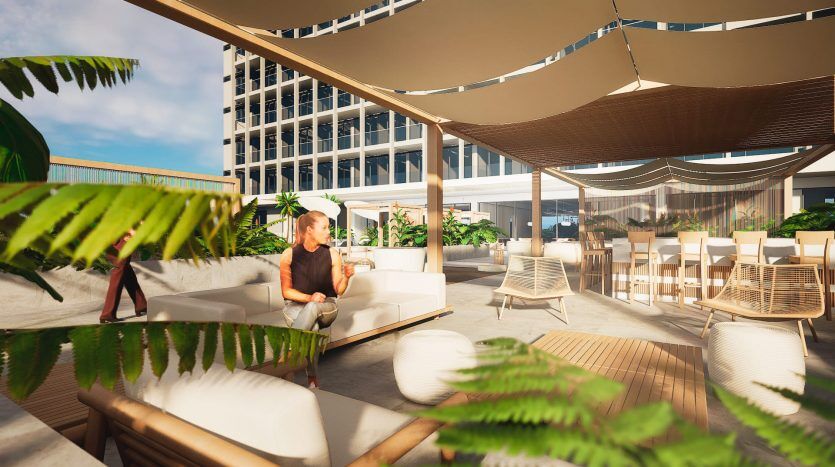 Une femme est assise seule sur un canapé blanc sous le patio ombragé d’une villa de Dubaï, entourée de plantes vertes luxuriantes avec des immeubles modernes de grande hauteur en arrière-plan. La lumière du soleil filtre à travers les stores en tissu