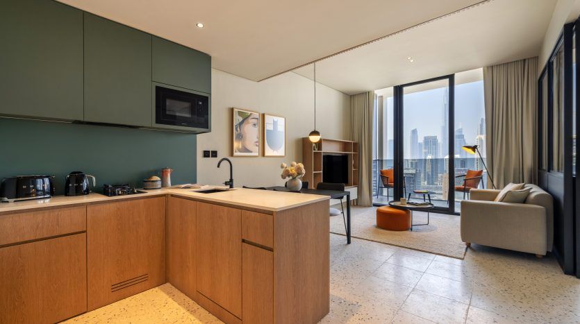 Cuisine moderne avec armoires en bois et murs verts, menant à un espace de vie doté de grandes fenêtres mettant en valeur un paysage urbain. Intérieur élégant, propre et lumineux d'un appartement à Dubaï.