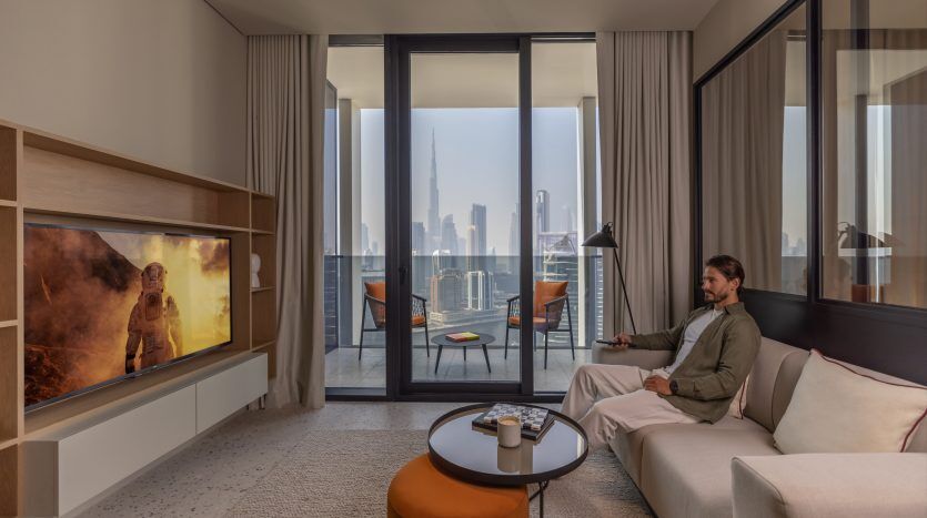 Un homme est assis détendu sur un canapé dans un salon moderne de son appartement de Dubaï, regardant un grand écran de télévision affichant une scène enflammée. La pièce offre une vue sur les toits de la ville du sol au sol.