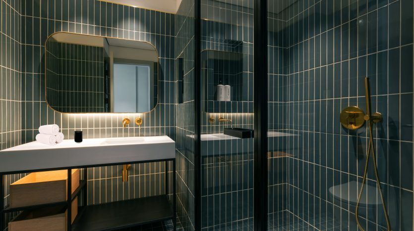 Une salle de bains moderne dans une villa de Dubaï avec des carreaux bleu foncé, un miroir rectangulaire, un lavabo blanc avec des luminaires dorés et un espace douche vitré avec une pomme de douche dorée.