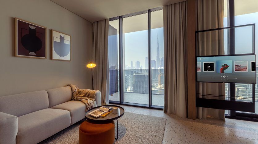 Salon moderne dans un appartement de Dubaï avec une grande fenêtre offrant une vue sur les toits de la ville, un canapé beige, une petite table ronde, un lampadaire, des rideaux et une télévision affichant une interface. Ouvrages d'art