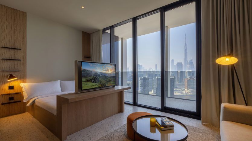 Une chambre d'hôtel moderne dotée d'un grand lit, d'une télévision à écran plat affichant une scène naturelle et d'une baie vitrée donnant sur les toits de Dubaï. La chambre comprend un mobilier élégant et un éclairage chaleureux.