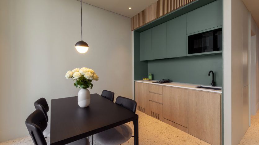 Une cuisine moderne dans un appartement de Dubaï au design minimaliste comprenant une table à manger sombre, des chaises noires et des armoires en bois clair avec des armoires supérieures vertes. Un simple vase à fleurs blanches orne la table sous