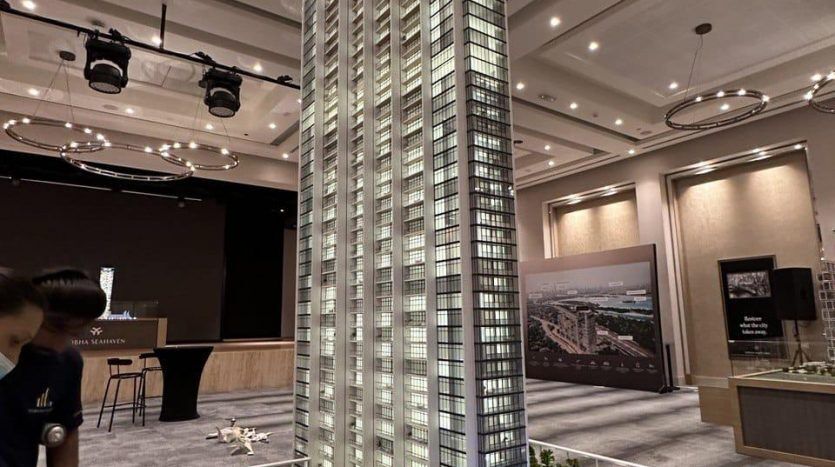 Un modèle architectural détaillé d'un grand gratte-ciel en verre placé sur une plate-forme surélevée, entouré d'arbres et de routes miniatures, exposé dans une grande salle avec des spectateurs observant, représentant un projet d'investissement immobilier de Dubaï.