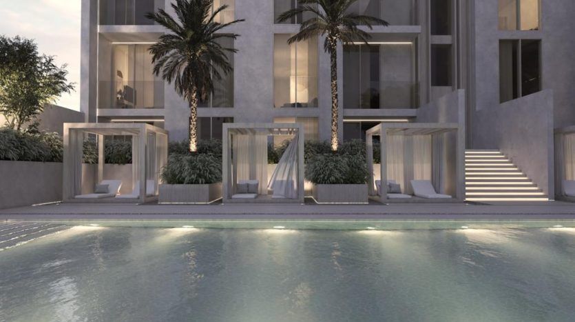 Maison à l'architecture moderne comprenant une piscine au premier plan avec des cabanes éclairées et des palmiers sur fond d'immeuble à plusieurs étages le soir, parfaite pour ceux qui s'intéressent à l'immobilier Dubaï.
