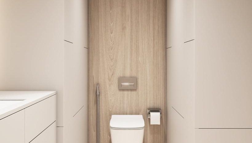 Une salle de bains moderne dans une villa à Dubaï comprenant des toilettes murales avec douchette de bidet, placées contre un panneau arrière en bois, flanquées de murs beiges doux et d'une vasque blanche sur la gauche.
