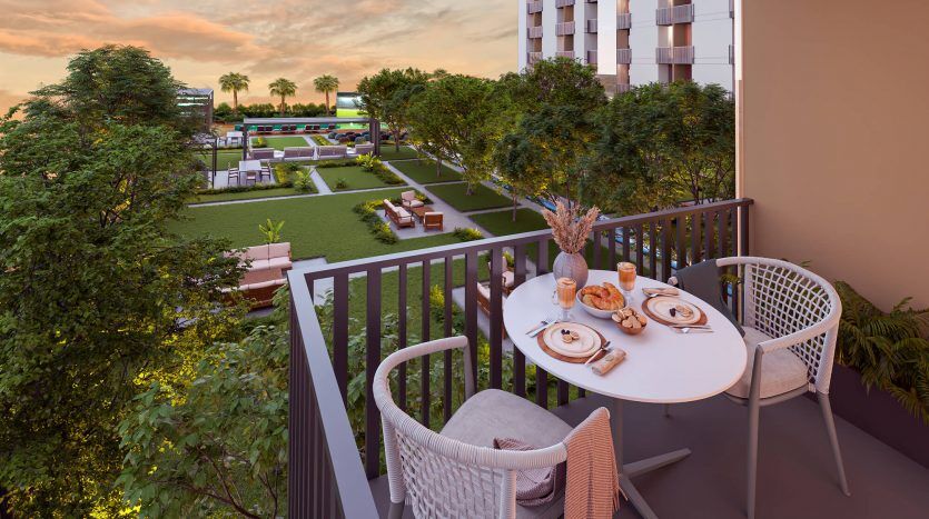 Un balcon avec vue sereine surplombant un jardin luxuriant au crépuscule, avec une petite table dressée pour deux, proposant un repas léger et des boissons, à côté d'un bâtiment urbain moderne à Dubaï.