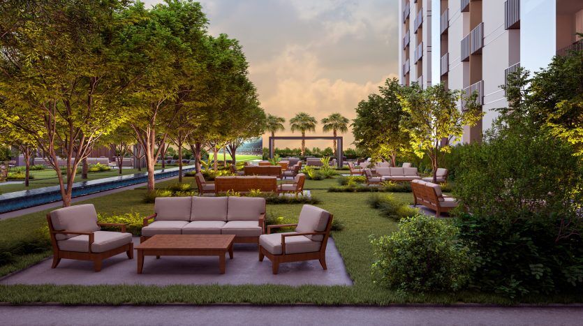 Un coin salon de jardin luxuriant entre des immeubles d'appartements modernes à Dubaï, doté de canapés et de fauteuils confortables, entouré d'arbres et de verdure, avec une pièce d'eau tranquille en arrière-plan sous un ciel couchant.