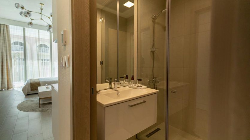 Un intérieur de salle de bains moderne comprenant une vanité blanche avec un lavabo, une cabine de douche en verre et un aperçu d'un espace de vie confortable à travers la porte, illustrant une opulente propriété immobilière de Dubaï.