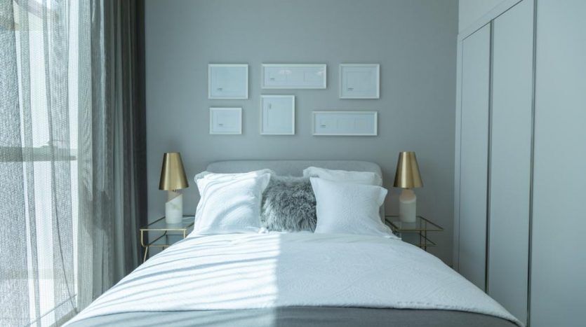 Une chambre moderne dans une villa à Dubaï comprenant un lit soigneusement fait avec une literie blanche et grise, flanqué de deux lampes dorées sur des tables de chevet à plateau en verre et décoré d'œuvres d'art encadrées au-dessus du lit.