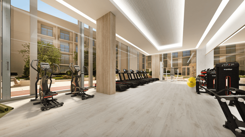 Une salle de sport spacieuse et moderne avec de grandes fenêtres, dotée de tapis roulants alignés, de machines elliptiques, de divers équipements de fitness et d'une ambiance lumineuse et accueillante idéale pour investir à Dubaï.