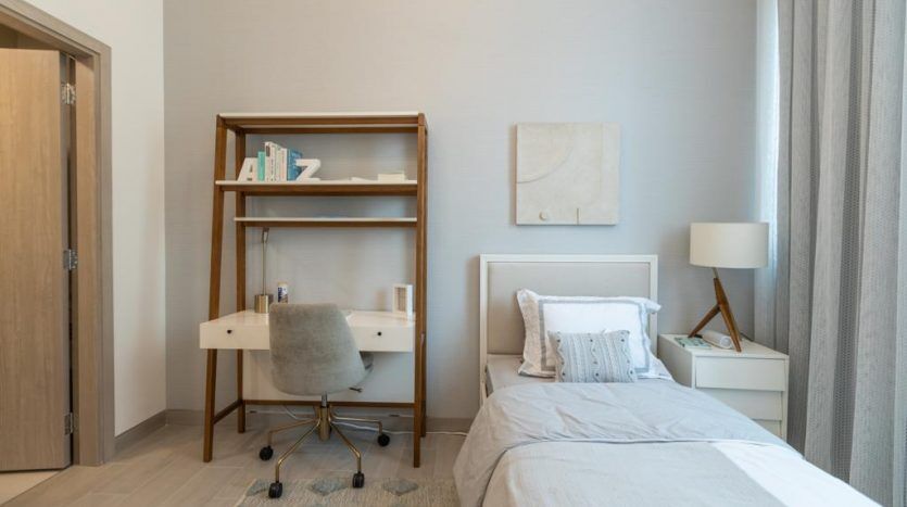 Une chambre moderne dans un appartement à Dubaï comprenant un lit simple avec une literie blanche, un bureau avec une chaise, une bibliothèque, des œuvres d'art murales abstraites et une lampe, le tout dans une palette de couleurs neutres apaisantes