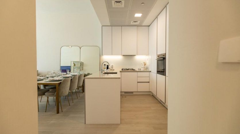 Une cuisine moderne avec des armoires blanches et des appareils électroménagers intégrés dans un appartement de Dubaï s'ouvre sur un coin repas avec une table dressée, vue depuis un couloir éclairé par une lumière chaleureuse.
