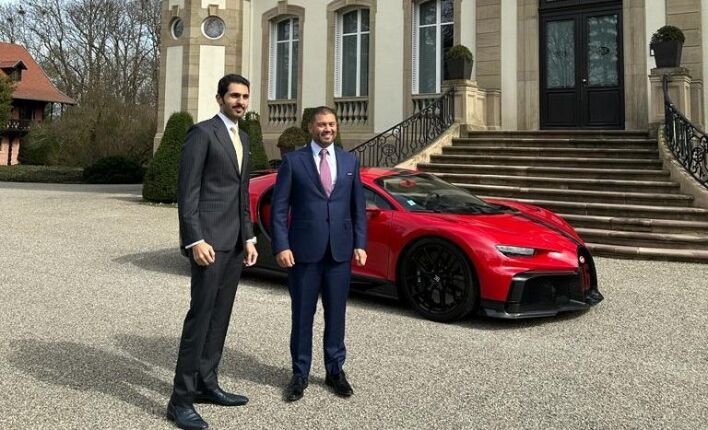 Deux hommes en costume d'affaires debout devant une voiture de sport rouge garée devant un élégant manoir à Dubaï avec un ciel clair en arrière-plan.