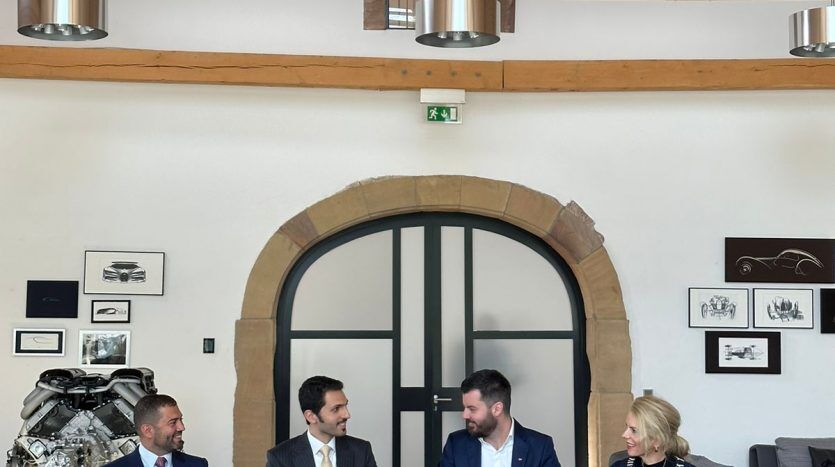 Quatre hommes d'affaires sont assis à une table dans une salle d'exposition moderne avec des poutres en bois apparentes et des pièces automobiles exposées autour d'eux. Une voiture de luxe est partiellement visible au premier plan, soulignant leur discussion sur les opportunités d'investissement dans