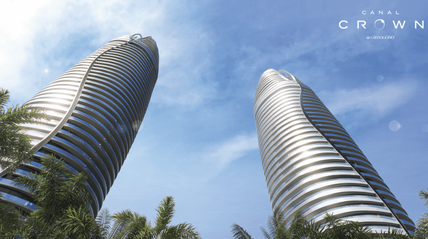 Deux gratte-ciel modernes et incurvés sous un ciel bleu clair avec des arbres verts luxuriants au premier plan et le logo « Canal Crown by Danzoo » au sommet, mettant en valeur le premier immobilier de Dubaï.