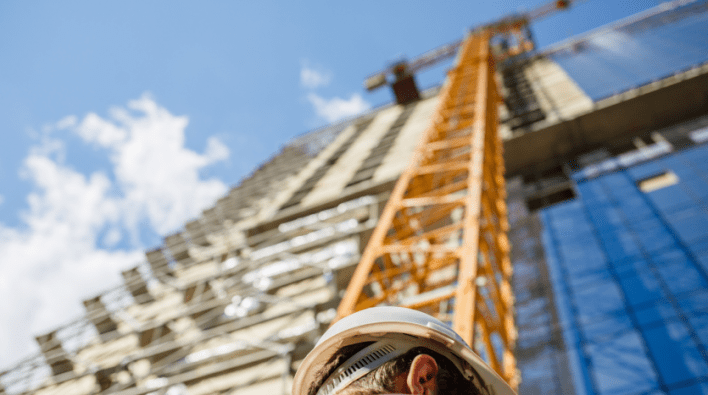 Chantier de construction à Dubaï avec un accent sur le casque d'un ouvrier au premier plan et une vue floue d'une grue et d'un gratte-ciel en construction en arrière-plan sous un ciel bleu.