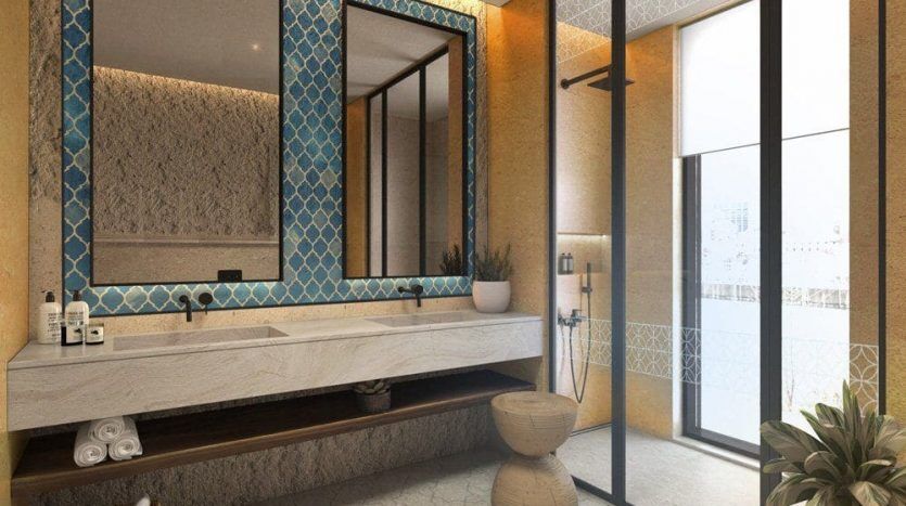 Un lujoso cuarto de baño en una villa de Dubai con paredes doradas, un gran espejo enmarcado en azul y oro, una encimera de madera, un lavabo y una puerta de ducha de cristal. Una planta y un tejido