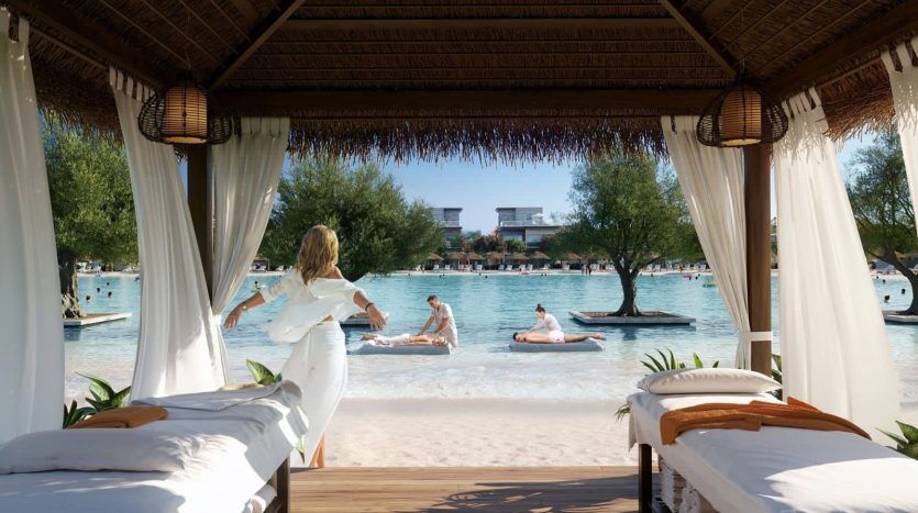 Una mujer vestida de blanco disfruta de las vistas desde una lujosa cabaña de playa con vistas a una tranquila laguna verde azulado donde la gente se relaja en tumbonas flotantes en Dubai.