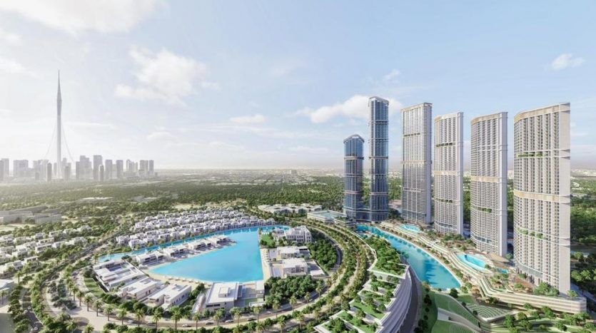 Aerial view of a modern urban development in Dubai with high-rise buildings, a large artificial lake and vast green spaces, under a clear sky.