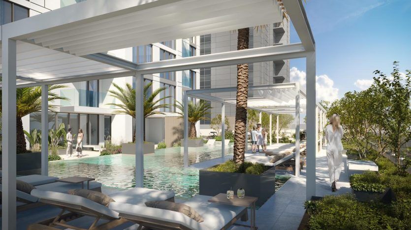 Luxurious outdoor pool area with modern loungers under a white pergola, surrounded by lush greenery and palm trees. People walk and relax casually in a sunny, serene setting, next to a glass building featuring the premier
