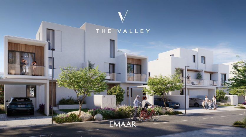 Rendu numérique d'un quartier résidentiel moderne de Dubaï avec des maisons de ville blanches contemporaines, mettant en vedette des personnes en interaction et des véhicules garés. Le logo de « emaar the valley » est affiché en haut.