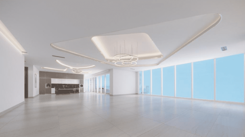Intérieur moderne et spacieux avec un vaste plancher, de grandes fenêtres offrant une lumière naturelle abondante, des plafonniers uniques et un espace de réception visible en arrière-plan. Cet appartement Dubaï présente un design minimaliste avec une couleur claire