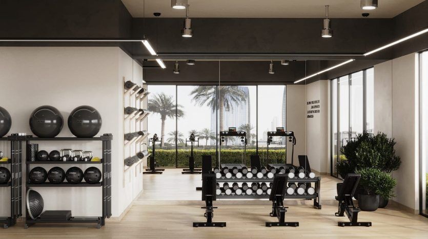 Un intérieur de salle de sport moderne avec des haltères, des bancs de musculation et des supports sous les plafonniers dans un appartement de Dubaï, donnant sur les palmiers à travers de grandes fenêtres en verre.