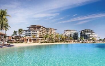 Complexe luxueux en bord de mer avec des bâtiments modernes à plusieurs étages, entouré de palmiers luxuriants, surplombant une grande piscine bleue sereine sous un ciel dégagé, idéal comme investissement immobilier à Dubaï.
