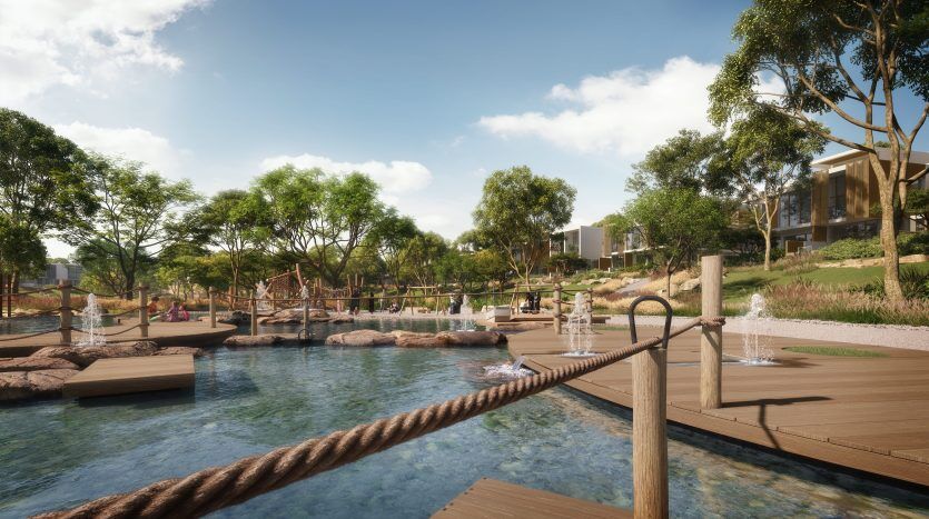 Un paysage serein au bord d'un lac avec des allées en bois, des balustrades en corde, entouré d'arbres luxuriants et des maisons modernes en arrière-plan sous un ciel clair, parfait pour investir à Dubaï.