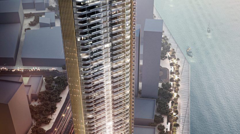 Un gratte-ciel rendu numériquement situé au bord de l'eau à Dubaï, présentant un extérieur élégant et doré avec un design ondulé, à côté d'un paysage urbain animé avec des bateaux dans l'eau.