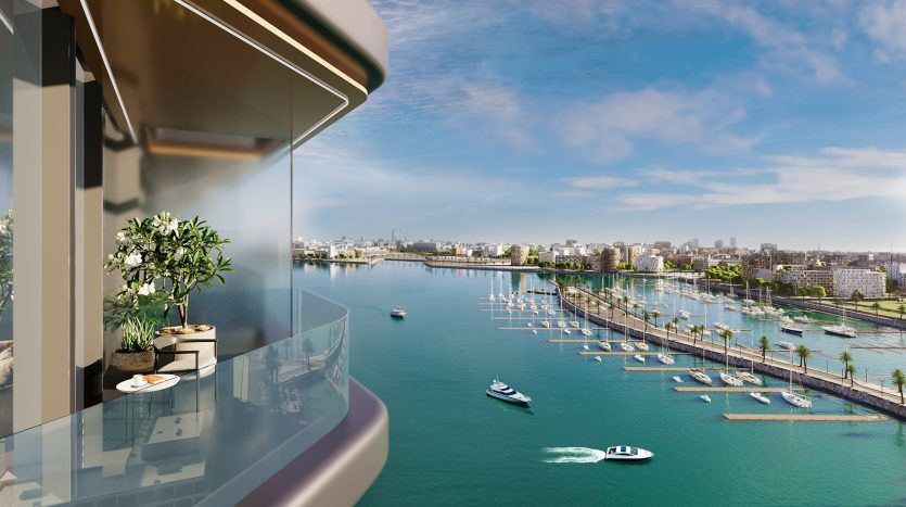 Une vue panoramique depuis un balcon en hauteur surplombant une rivière animée de bateaux. Une silhouette de ville remplit l’horizon sous un ciel clair. Des plantes en pot décorent le balcon de l'appartement Dubaï.