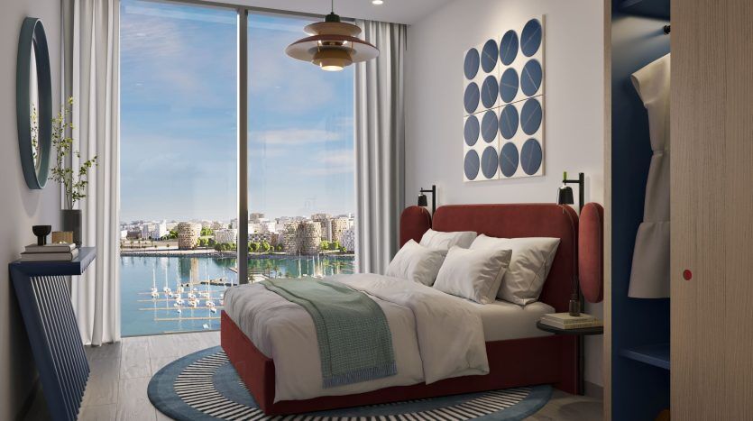 Chambre moderne dans un appartement de Dubaï avec une grande fenêtre donnant sur un paysage urbain et sur l'eau. Comprend un lit rouge avec une literie blanche, un plafonnier rond, des touches bleues et une décoration minimaliste.