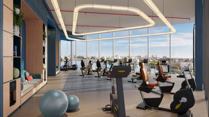 Salle de sport moderne avec divers équipements d'entraînement, notamment des tapis de course et des vélos, donnant sur un paysage urbain à travers de grandes fenêtres. L'intérieur présente des accents en bois et des plantes vertes, parfaits pour investir dans l'immobilier axé sur la santé de Dubaï.