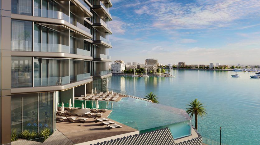 Luxueux immeuble de grande hauteur en bord de mer à Dubaï avec balcons donnant sur une marina remplie de bateaux, doté d'une piscine à débordement et de transats.