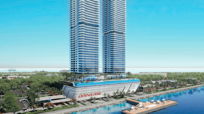 Vue aérienne d'un gratte-ciel moderne au bord de l'eau à Dubaï, affichant le logo « Danube » à sa base. Des arbres et une marina avec des bateaux sont visibles le long du rivage.