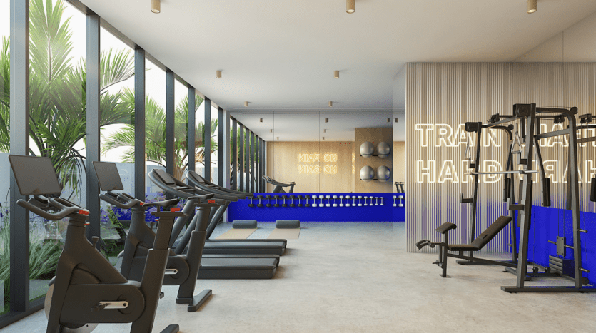 Intérieur de salle de sport moderne comprenant des tapis roulants, des bancs de musculation et divers appareils d'exercice, avec de grandes fenêtres présentant des palmiers et de la lumière naturelle. Les murs sont ornés d’enseignes lumineuses motivantes. Parfaitement positionné pour ceux