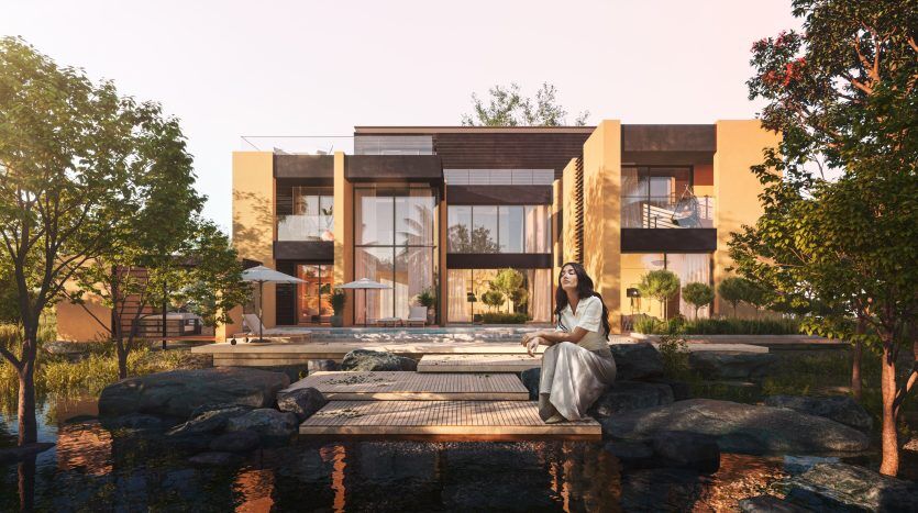Une femme est assise paisiblement sur une terrasse en bois au bord d'un étang tranquille devant une luxueuse et grande villa en verre et en bois à Dubaï, entourée d'une verdure luxuriante au coucher du soleil.