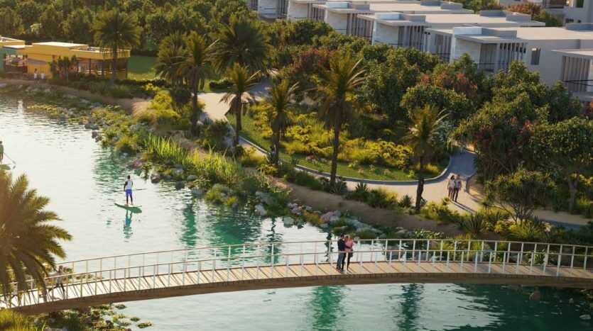 Une vue panoramique sur un parc luxuriant avec des gens marchant et faisant du paddleboard le long d'une rivière, avec un pont incurvé et des immeubles d'appartements modernes à Dubaï en arrière-plan.